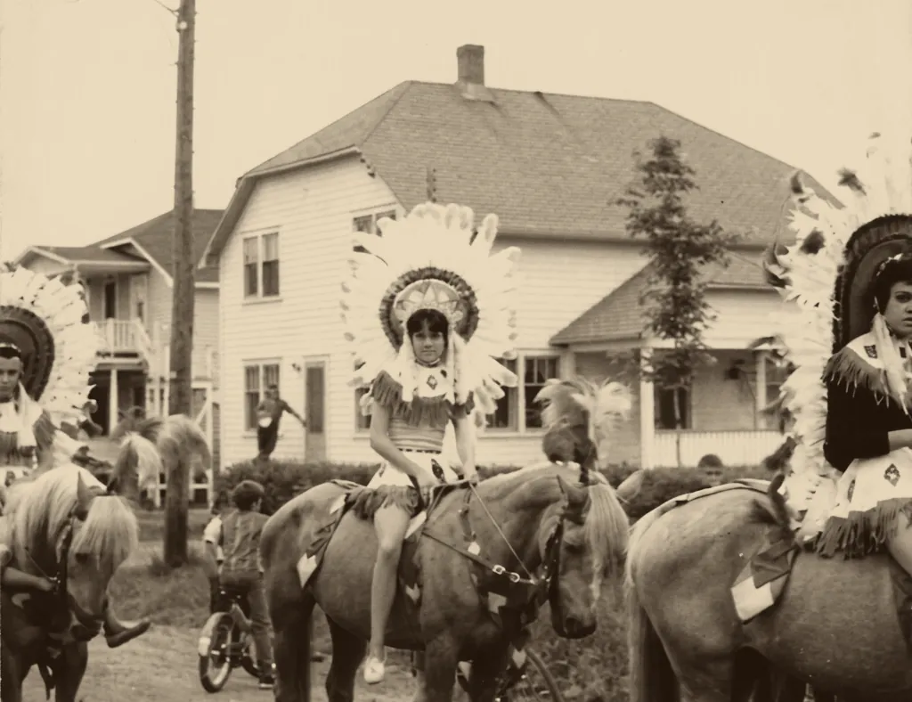 Garden Bros Circus in the 1930s: young riders in feathered headdresses on horseback in front of a white house